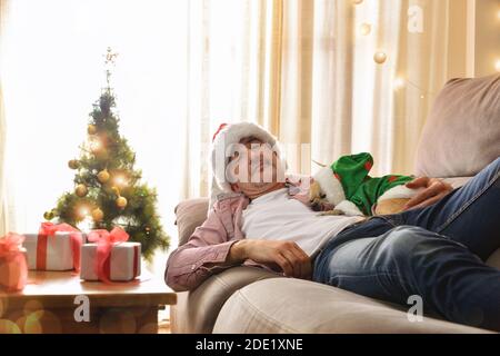 L'uomo che dorme con il suo cane alle feste di Natale su un divano in un vestito noel con sfondo decorato con albero e regali. Foto Stock