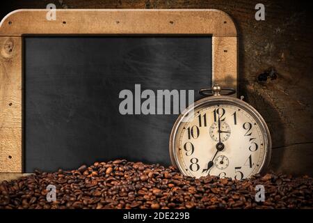 Closeup di una vecchia sveglia e un mucchio di chicchi di caffè arrosto su una lavagna vuota con cornice in legno e spazio per la copia. Foto Stock