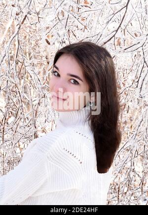 Ritratto di giovane donna russa in maglione bianco guardando la macchina fotografica accanto a rami congelati di alberi di betulla, coperto di ghiaccio. Foresta invernale in Russia Foto Stock