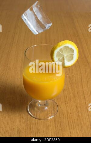 Un succo naturale di mela e arancia appena spremuto sul tavolo di legno in una cucina rustica con un cubetto di ghiaccio che cade nella bocca del bicchiere. Foto Stock