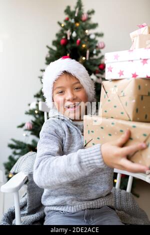 Ragazzo in cappello di clausola di santa davanti a un Albero di Natale con regali di Natale Foto Stock