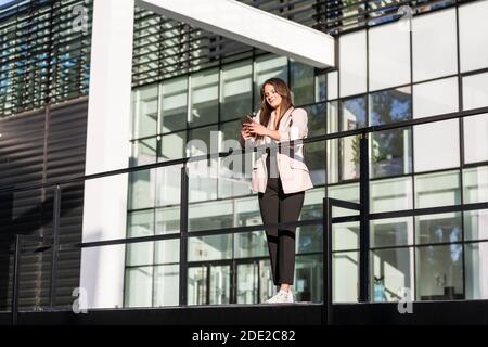 Una giovane bella donna appoggiata contro una recinzione e sorridente mentre scrive sul suo telefono cellulare. È addestrata al business, con architettura moderna nella b Foto Stock