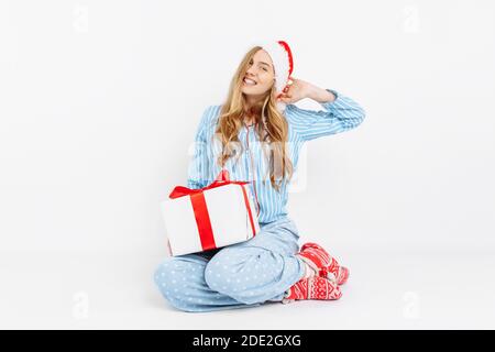 Mattina di Natale. Felice bella giovane ragazza, in pigiama di Natale e cappello di Santa, apre regalo di Natale, su sfondo bianco Foto Stock