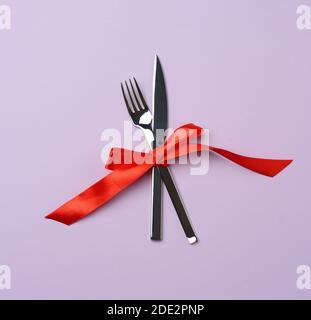 forchetta e coltello in metallo legati da un nastro di seta rossa, sfondo viola, vista dall'alto Foto Stock
