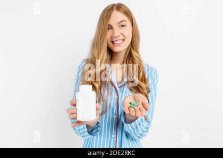 Una ragazza in pigiama contiene un vaso di plastica bianco per pillola, un concetto per il trattamento medico delle malattie, un primo piano di un vaso bianco per pillola, su uno sfondo bianco Foto Stock