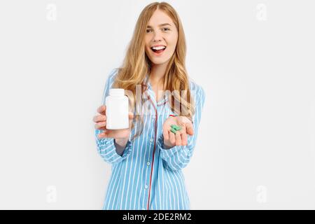 Una ragazza in pigiama contiene un vaso di plastica bianco per pillola, un concetto per il trattamento medico delle malattie, un primo piano di un vaso bianco per pillola, su uno sfondo bianco Foto Stock