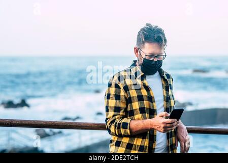 Ritratto di uomo adulto all'aperto con maschera di protezione del viso per coronavirus covid-19 focolaio - persone con maschera di protezione che guarda il telefono per notizie Foto Stock