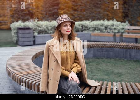 Affascinante giovane donna francese in un cappotto beige autunno e cappello plaid si siede su banco rotondo fiore letto in attesa per la sua data o fidanzate. Foto colorata Foto Stock