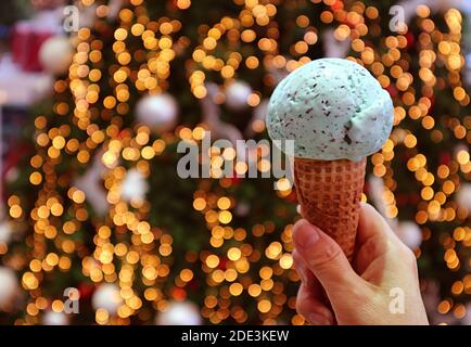 Primo piano di una tazza di cono gelato al cioccolato con chip di menta In mano con l'albero di Natale scintillante Bokeh in background Foto Stock