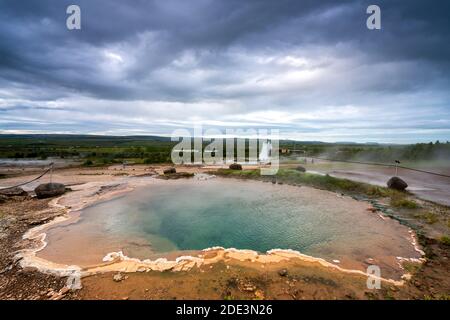 Vista panoramica del laghetto blu sulla zona geotermica con il geyser Strokkur che erutta sullo sfondo, Geysir, Islanda Foto Stock