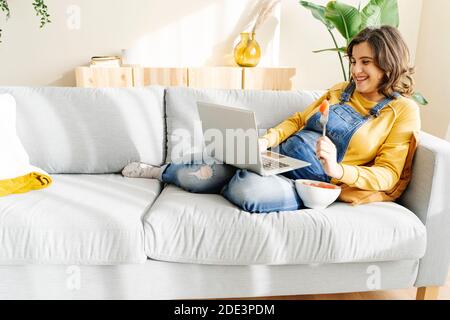 Sorridente giovane donna incinta che lavora sul suo portatile e mangia frutta su un divano. concetto di lavoro a casa Foto Stock