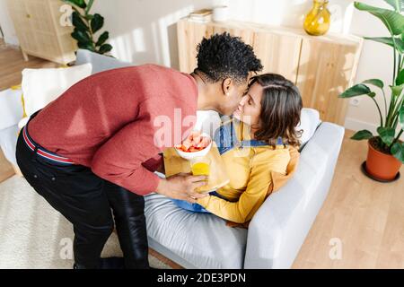 Marito che porta la colazione alla moglie incinta sul divano. Concetto di coppia interrazziale Foto Stock