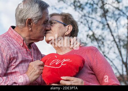 Vista laterale ad angolo basso di una romantica coppia con Cuore rosso abbracciando e baciando dolcemente mentre festeggia l'anniversario di nozze Foto Stock