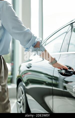 Mano di giovane donna elegante in casualwear in piedi da lussuoso auto nera e tenuta dalla maniglia mentre si apre una delle porte Foto Stock