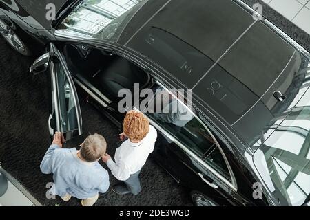 Vista di un giovane venditore di auto centro di apertura della porta di veicolo nero mentre mostra e descrive le caratteristiche della vettura interno al cliente maschile Foto Stock