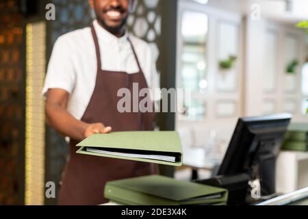 Mano di giovane cameriere felice in grembiule marrone che vi passa menu in copertina verde mentre si è in piedi accanto al suo posto di lavoro con ristorante Foto Stock