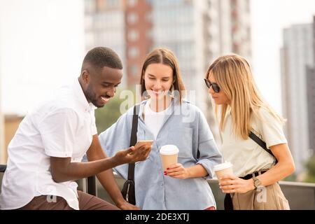Felici giovani donne bionde e brunette in casualwear che beve mentre si guarda il display dello smartphone in mano a bello Ragazzo africano Foto Stock