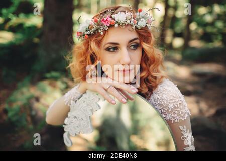 bella giovane sposa dai capelli rossi nella foresta con una corona floreale sulla testa e specchio. donna in lungo vestito bianco all'aperto il giorno d'estate. matrimonio Foto Stock