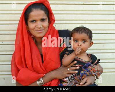 Giovane felice indiana Gujarati Rabari donna indossa una sciarpa rossa della testa della dupatta e tiene la sua carina bambina succhiata con il pollice tra le braccia. Foto Stock