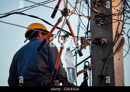 Lineman elettrico in un elmetto giallo che lavora con morsetti in altezza sui cavi delle strade, sopra un palo fissato da cinture Foto Stock