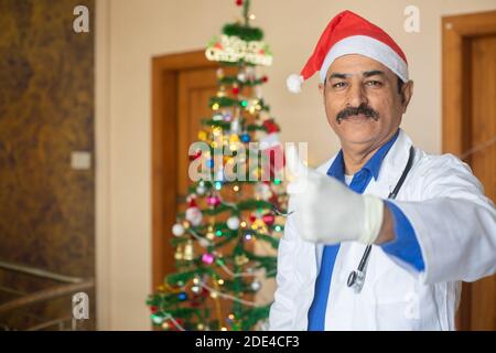 Medico di mezza età che indossa il cappello di natale facendo il pollice in su, faccia felice dell'uomo sorridente. Celebrazione durante la pandemia di covid-19, blocco, soggiorno a casa, nuova norma Foto Stock