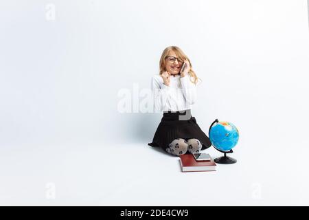 Una bambina, a immagine di un insegnante, indossando occhiali con un globo e libri, parlando al telefono e gioendo e ridendo, su una schiena isolata Foto Stock