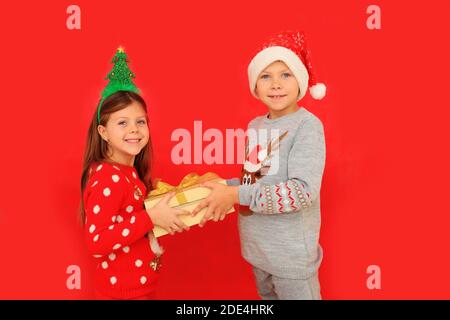 Due bambini in pullover hanno in mano un regalo di Capodanno su sfondo rosso. Un ragazzo e una ragazza danno un regalo di Natale a vicenda Foto Stock