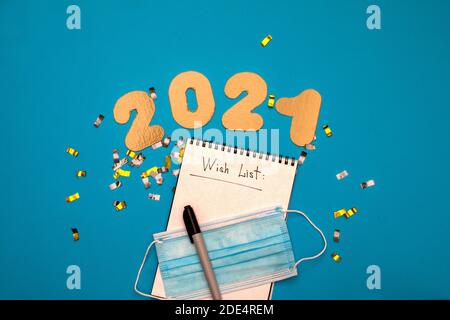 Vista dall'alto del pennarello e della maschera medica taccuino con lista dei desideri vicino a confetti e 2021 iscrizioni sopra sfondo blu Foto Stock