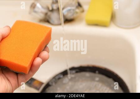 spugna per lavare piatti in mano a un popolo sullo sfondo del lavandino della cucina con un sporco vecchia padella Foto Stock