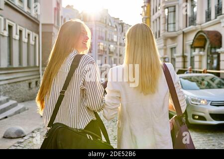 Due giovani donne con borse per computer portatili che camminano lungo la strada della città del tramonto, vista posteriore