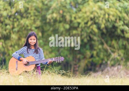 Una carina studentessa asiatica dai capelli lunghi suonava felicemente la chitarra in erba al mattino. Foto Stock