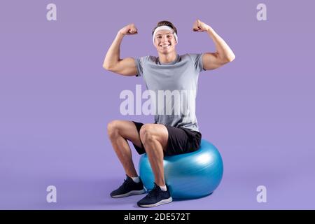 Positivo giovane sportivo seduto su palla fitness e mostrandogli forza su sfondo lilla studio Foto Stock