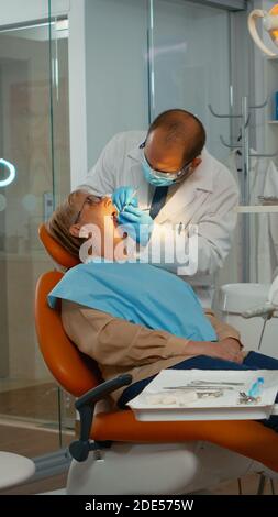 Medico che esamina i denti con strumenti medici che lavorano con i guanti in una moderna clinica dentista. Ortodontista che parla con la donna seduta sulla sedia stomatologica mentre l'infermiere prepara gli attrezzi per l'esame Foto Stock