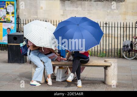 Cambridge, UK, 1 agosto 2019. Ragazze seduta sul banco di nascondersi dietro gli ombrelloni Foto Stock