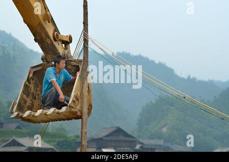 ZHAOXING DONG, Cina - 21 giugno 2012: Cinese lavoratore esterno di fissaggio cavi di energia elettrica a traliccio. Egli è seduto all'interno dell'escavatore a benna. La minoranza Miao Foto Stock