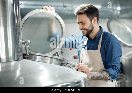 Sorridente lavoratore maschio o proprietario in grembiule apre coperchio di grande caldaia per la preparazione della birra e il controllo della fermentazione Foto Stock