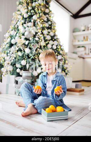 Bambino piccolo in pigiama blu si siede a piedi nudi sul pavimento caldo e tiene una scatola di tangerini sullo sfondo di un albero di Natale. Foto Stock