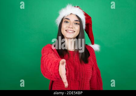 Giovane bella donna che indossa il cappello di Natale su verde isolato sfondo sorridente amichevole offrendo handshake come saluto e accogliente Foto Stock