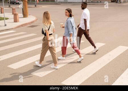 Tre giovani amici interculturali felici in casualwear che beve e. parlare mentre si attraversa la strada e si cammina verso l'altro lato di via Foto Stock