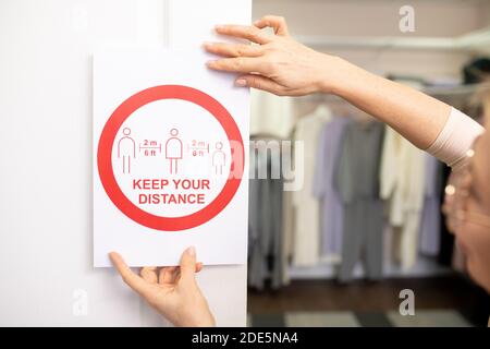 Mani di assistente maturo del negozio che mette la scheda che dice circa mantenere distanza a parete contro il rack con nuova raccolta di calore abiti e pullover Foto Stock