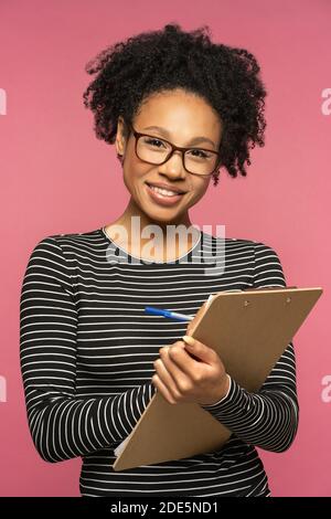 Giovane insegnante afro-americana felice isolata sulla parete rosa dello studio. La ragazza studentesca indossa gli occhiali che tengono la cartella, sorridente, guardando la macchina fotografica. Istruzione Foto Stock