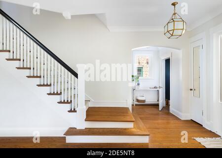 Una scalinata rinnovata nel foyer della casa con vista di un fangio e porta ad un piccolo bagno blu. Una luce moderna pende sopra i pavimenti in legno. Foto Stock