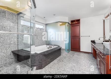 Un bagno grande e moderno con piastrelle di marmo grigio e bianco sul pavimento e sulle pareti con una doccia di vetro, vasca idromassaggio e armadietti in legno. Foto Stock
