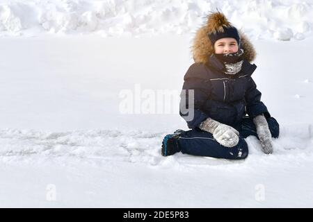 Il bambino si siede sul ghiaccio coperto di neve, circondato da nevicate. Vestiti in abiti invernali, leggermente nevosi. In una giornata limpida e soleggiata. Foto Stock