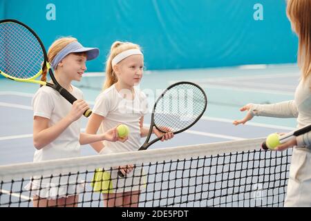 Due ragazze carine con racchette da tennis che guardano una giovane femmina trainer mentre si è in piedi dalla rete davanti a lei e. ascoltare le spiegazioni Foto Stock