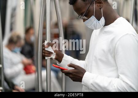 Afro American uomo indossare maschera viso nel trasporto pubblico durante la pandemia covid-19, coronavirus, tiene il corrimano attraverso il tovagliolo, utilizzando mobili s. Foto Stock