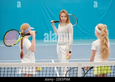 Giovane allenatore di tennis femminile sicuro in activewear che tiene la racchetta dietro collo e guardando due ragazze mentre le consultano sopra stadio Foto Stock