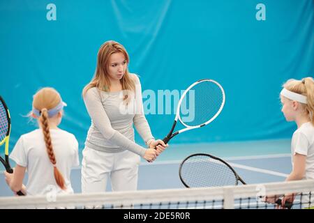 Giovane allenatore di tennis femminile bionda in activewear tenuta racchetta pronta spingere la palla e guardando due ragazze mentre consultandosi loro sullo stadio Foto Stock