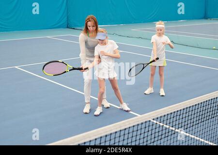 Giovane allenatore di tennis in Activewear tenuta racchetta da uno di due ragazze carine mentre consultantela durante l'addestramento individuale sullo stadio Foto Stock