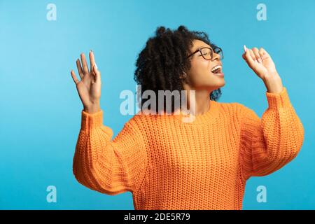 Donna millennial con pelle scura divertente e capelli ricci di buon umore canta la sua canzone preferita, ballando, indossa hipster oversize maglia arancione, isolato su s. Foto Stock